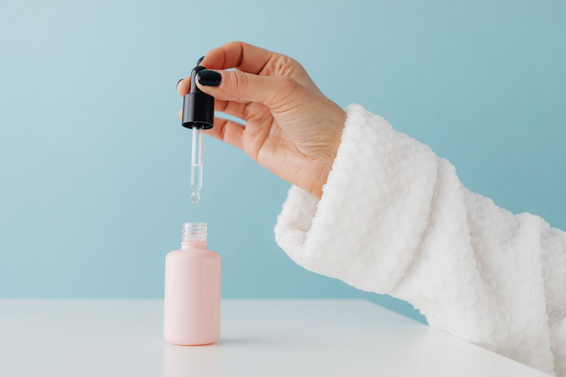 Close-up of a hand holding a glass dropper above a serum bottle on a neutral background