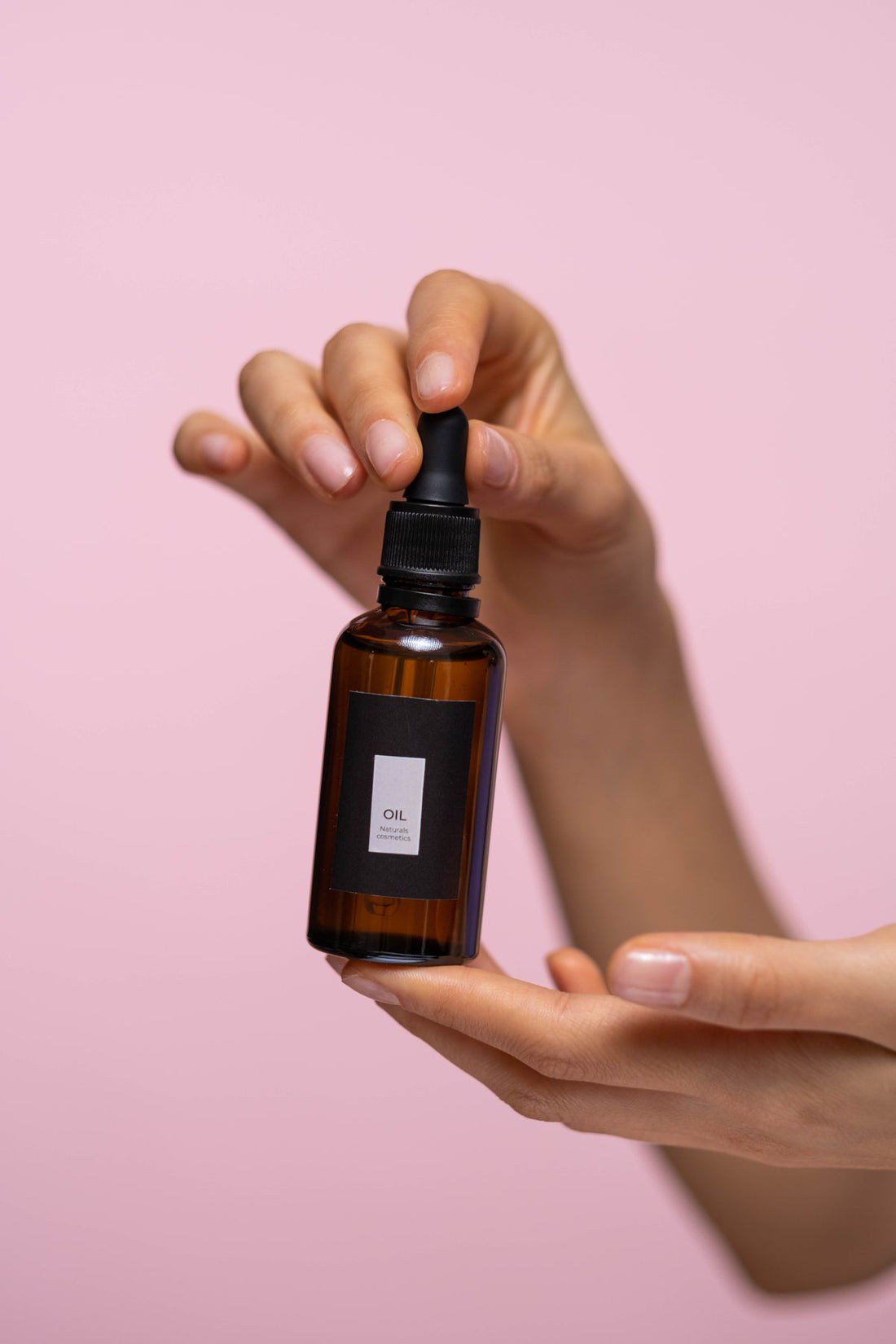 Close-up of hands holding a brown glass dropper bottle of face serum against a soft pink background.